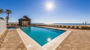 A rectangular swimming pool with clear blue water, surrounded by a tiled deck with lounge chairs. A small building with a sloped roof is adjacent to the pool. - Romar Place 104 |  Beachfront Condo | Indoor & Outdoor Pools | Beach Getaways (Orange Beach)