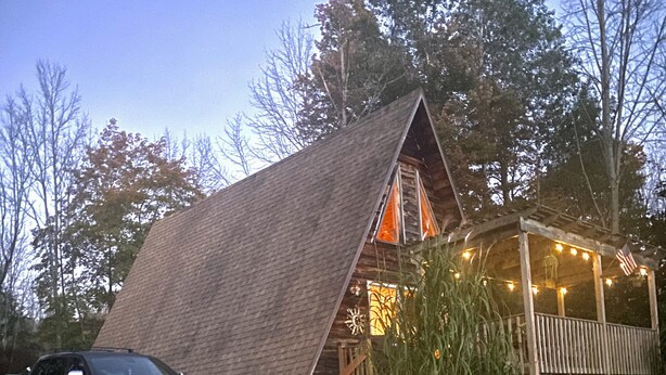 Una cabaña de madera con techo inclinado, un porche con luces de cuerda y un coche estacionado adelante.