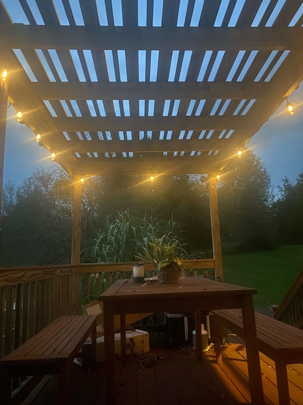 Puinen terassi, jonka yläpuolella on pergola, ja jossa on valonauhoja, pöytä ja tuoleja.