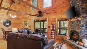 A cabin interior with a stone fireplace, leather sofa, dining area, and large windows. - Dog-Friendly! Secluded Cabin! Only 10 mins to downtown! Hot Tub & Game Room (Blue Ridge)