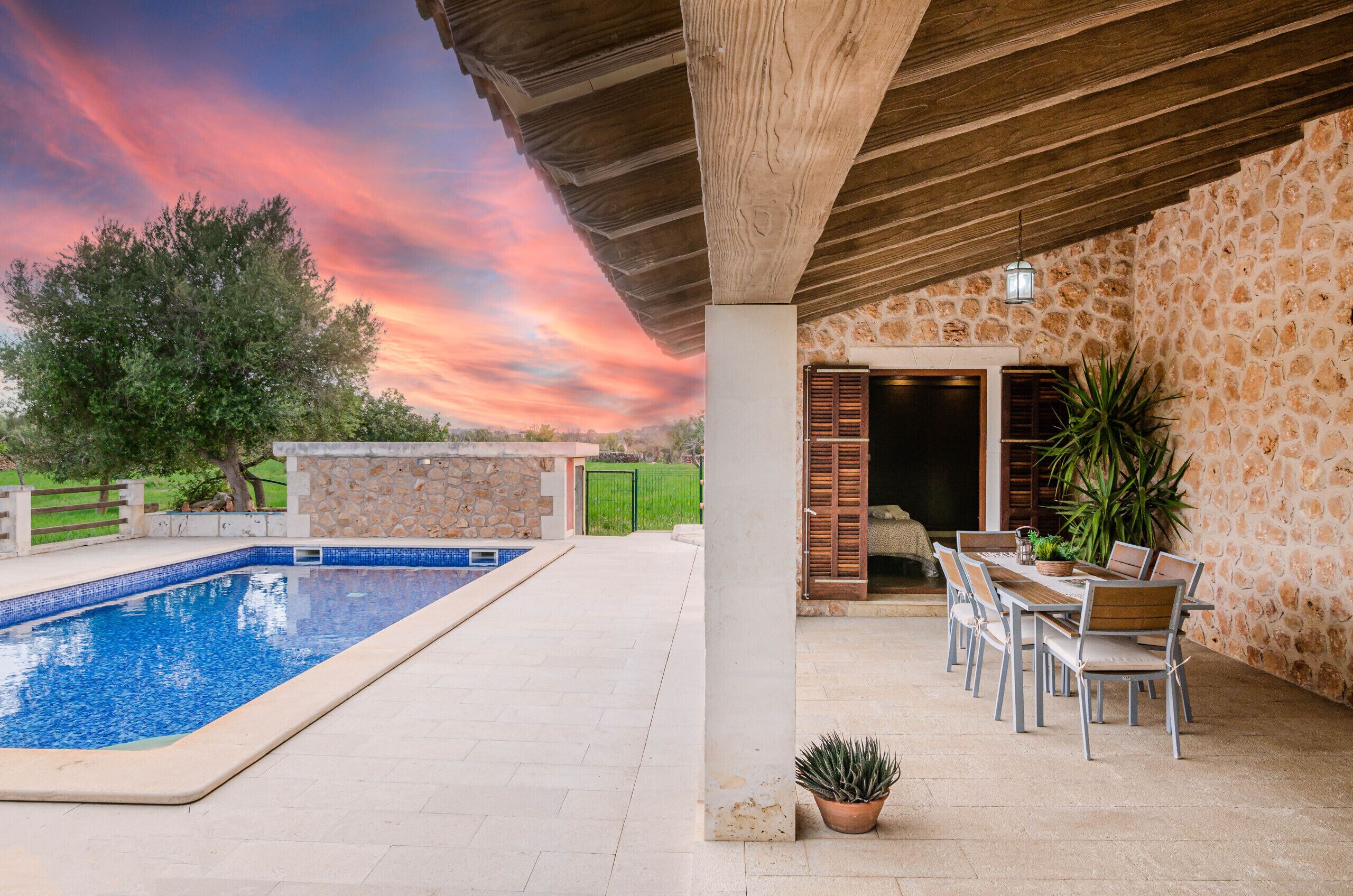 A pool area with a stone wall and wooden ceiling, a dining table with chairs, and a potted plant.