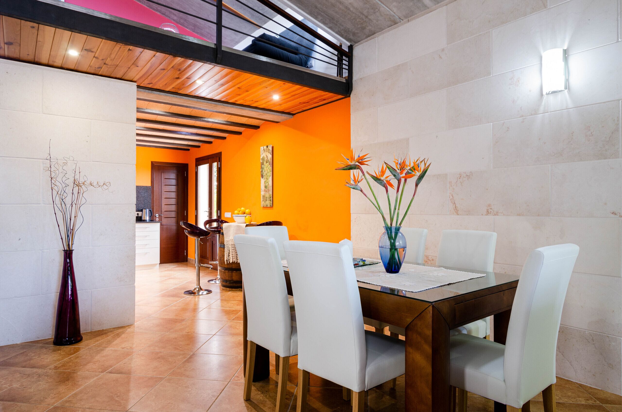 A dining area with a wooden table and white chairs, a vase with flowers, and a kitchen in the background.