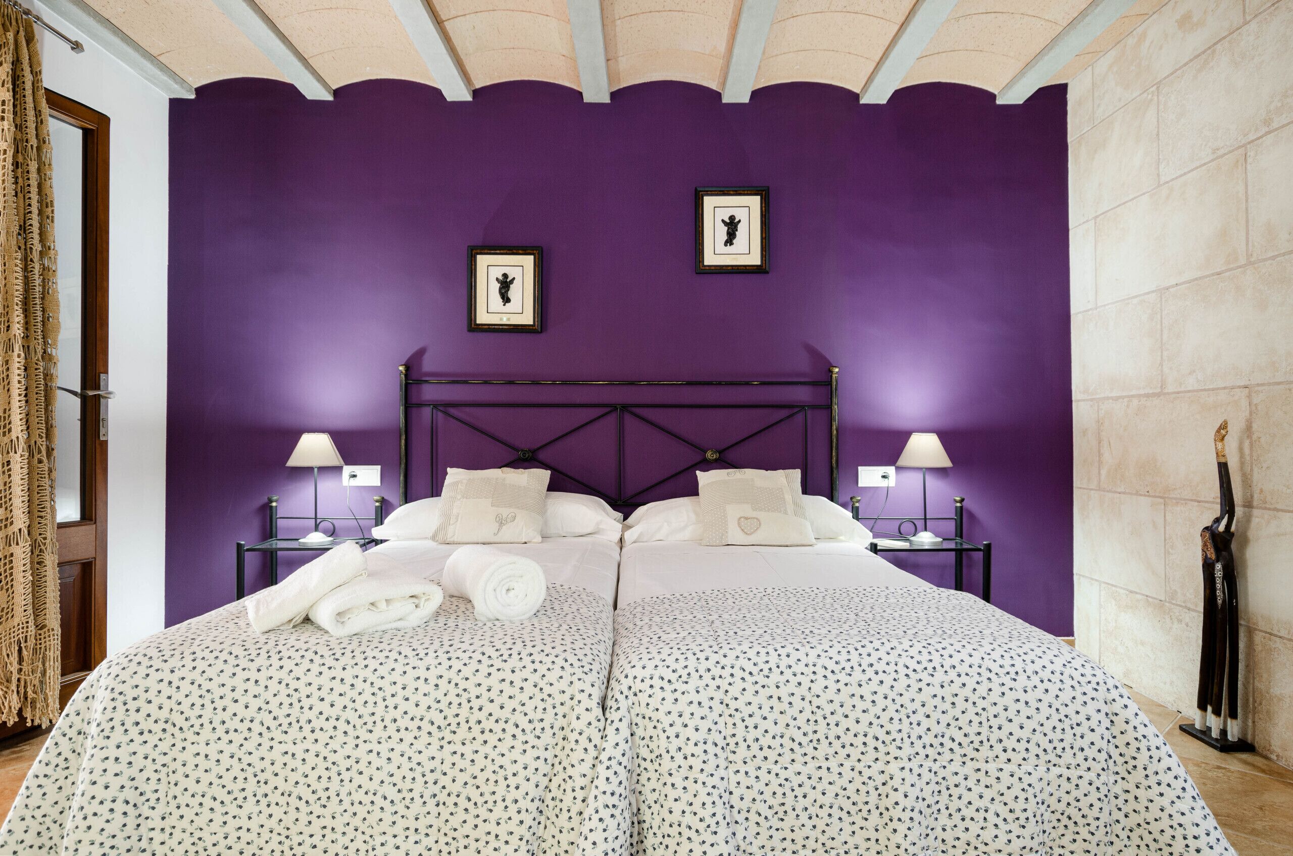 A bedroom with a purple wall, a bed with white linens, two framed pictures on the wall, and a nightstand with a lamp.