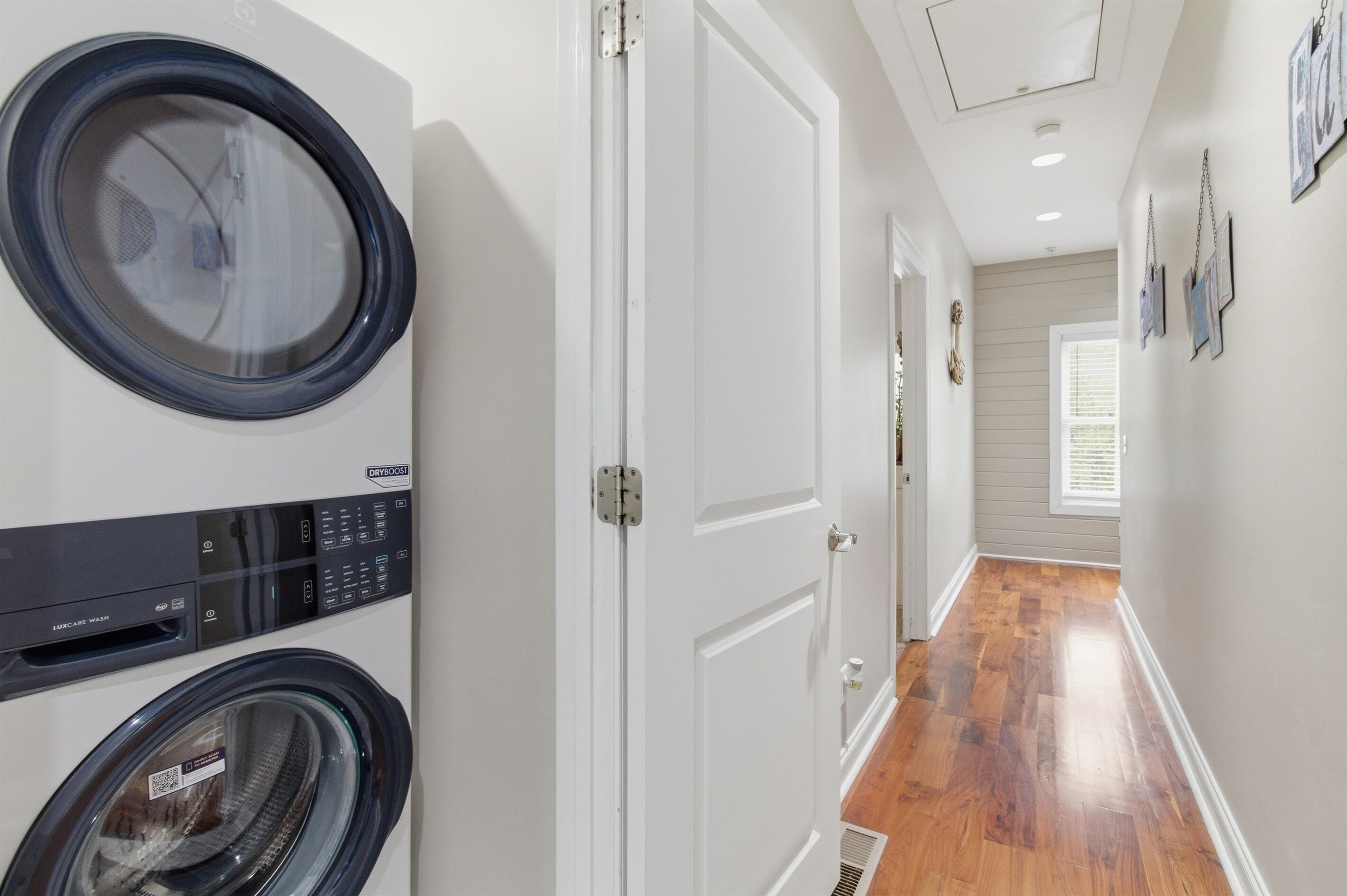 A laundry room with a front-loading washer and dryer, a white door, and a hallway with wooden flooring.