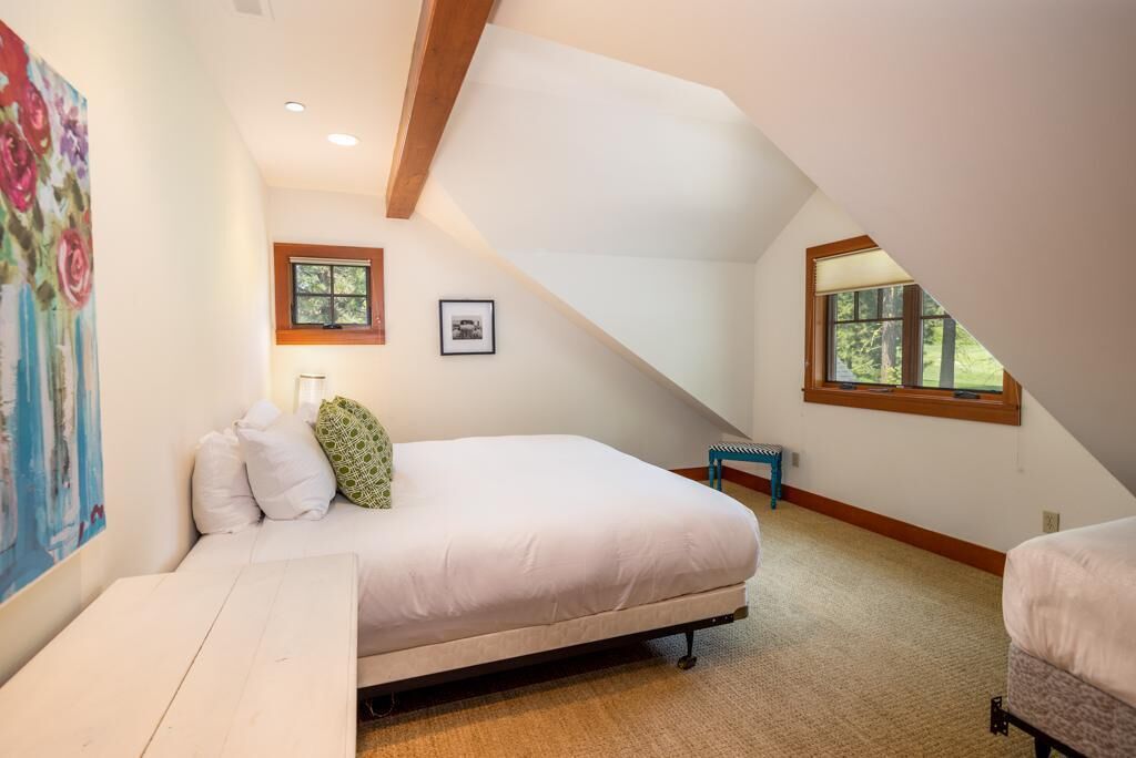 A bedroom with a loft bed, a small table, a bench, and a window with a view of trees.