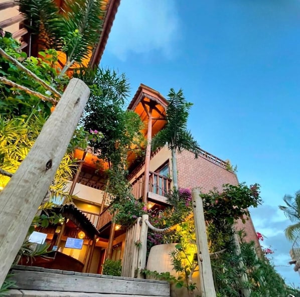Un edificio de varios pisos con balcones, rodeado de abundante vegetación y una valla de madera.