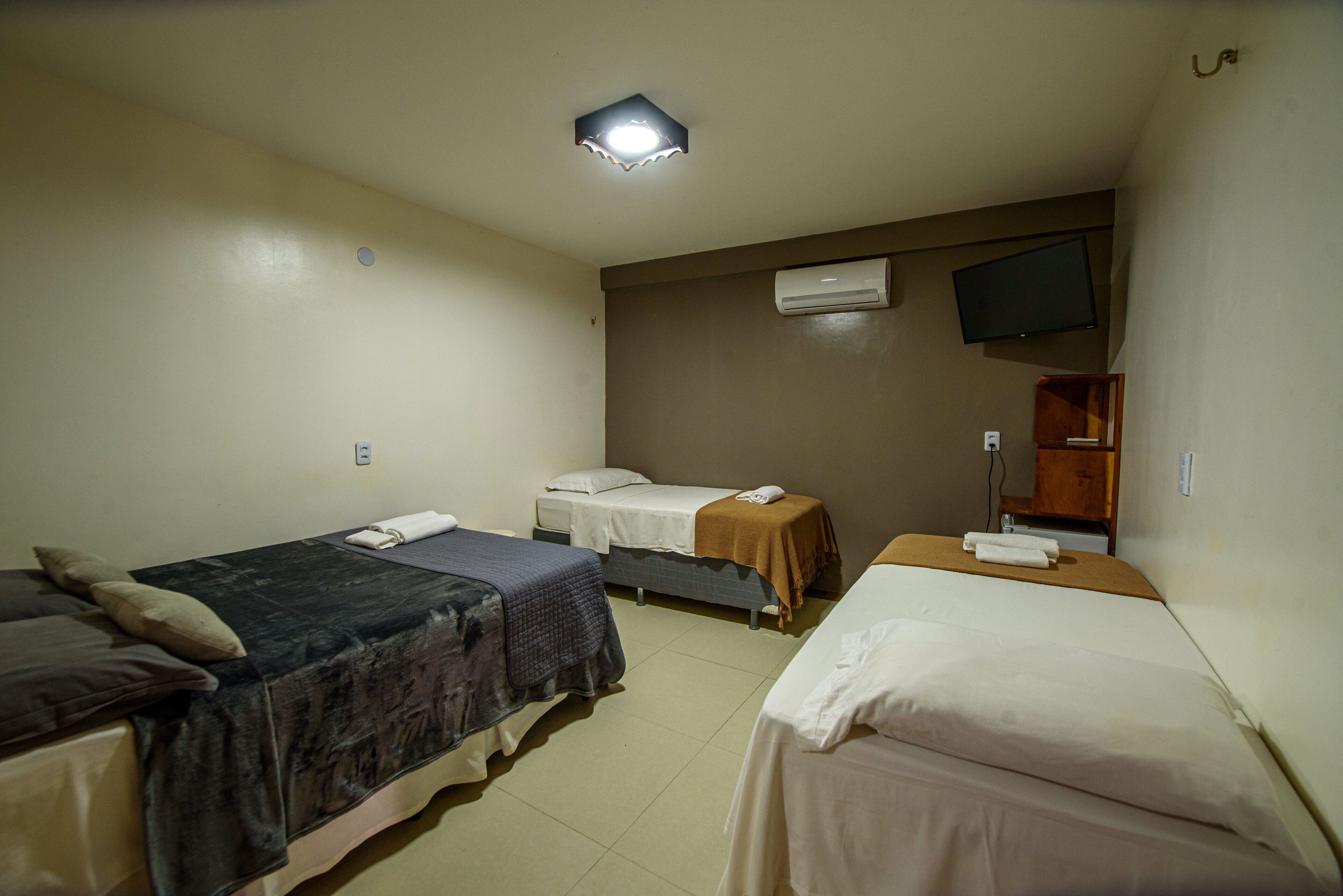 Habitación de hotel con tres camas individuales, un televisor de pantalla plana en la pared, un equipo de aire acondicionado y un pequeño armario de madera.