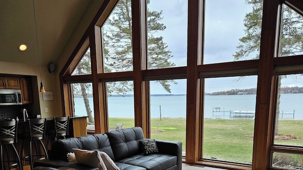 Un vaste salon doté d’une grande fenêtre donnant sur un lac, d’un confortable canapé en cuir noir et d’un coin cuisine avec des meubles en bois et une table à manger.