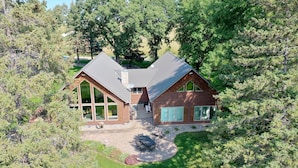 Une cabane en rondins à deux étages, dotée d’une terrasse en pierre et d’une grande fenêtre donnant sur une zone boisée.