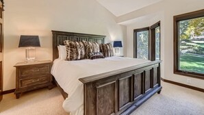 A bedroom with a large bed, a nightstand with a lamp, and a window with a view of trees. - 1-10 N. Fairway Drive (Avon)