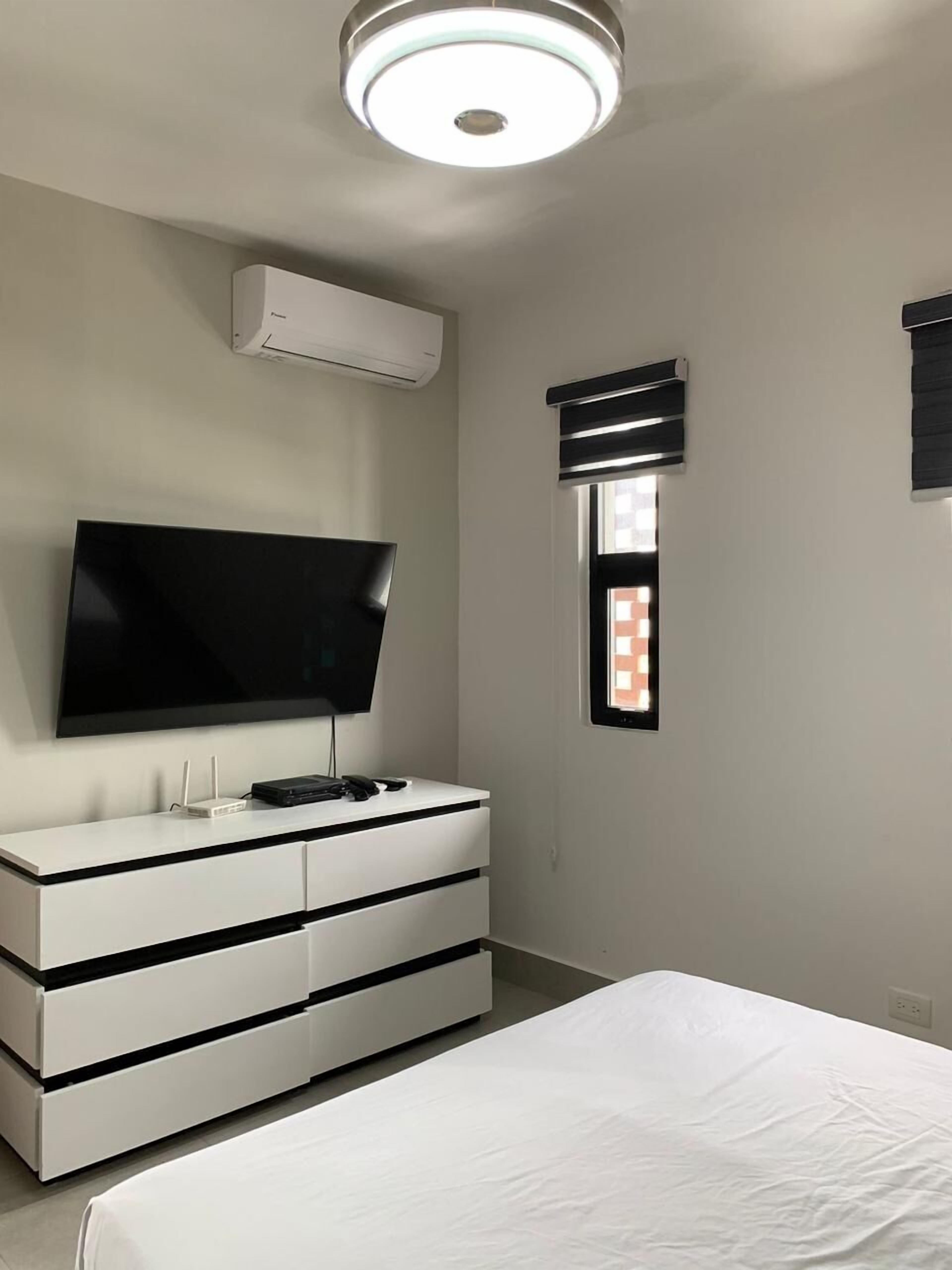 A bedroom with a bed, a TV mounted on a dresser, an air conditioning unit, and a window with blinds.