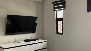 A bedroom with a bed, a TV mounted on a dresser, an air conditioning unit, and a window with blinds.