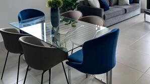 A modern living room with a glass dining table, blue chairs, a grey sofa, and a large window with blinds. - Beautiful view, 13th floor, modern, exclusive and comfortable apartment. (Santiago De Los Caballeros)