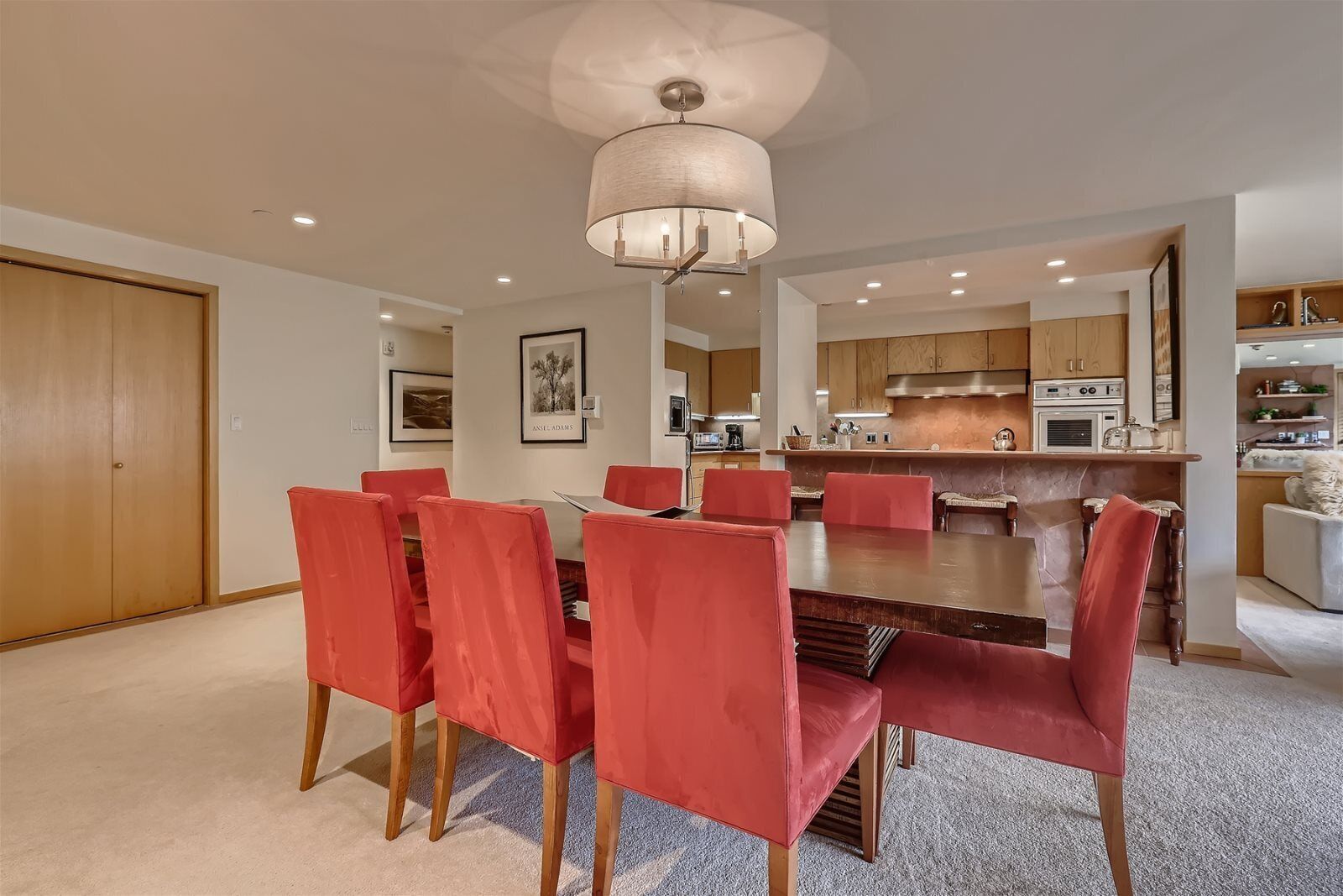 A modern dining area with a wooden table and red chairs, a kitchen with wooden cabinets, and a ceiling light fixture.
