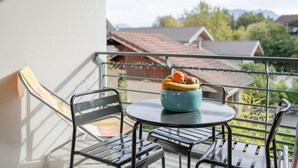 Ein Balkon mit rundem Tisch und zwei Stühlen, eine blaue Schale mit Früchten und Blick auf Häuser und Bäume.