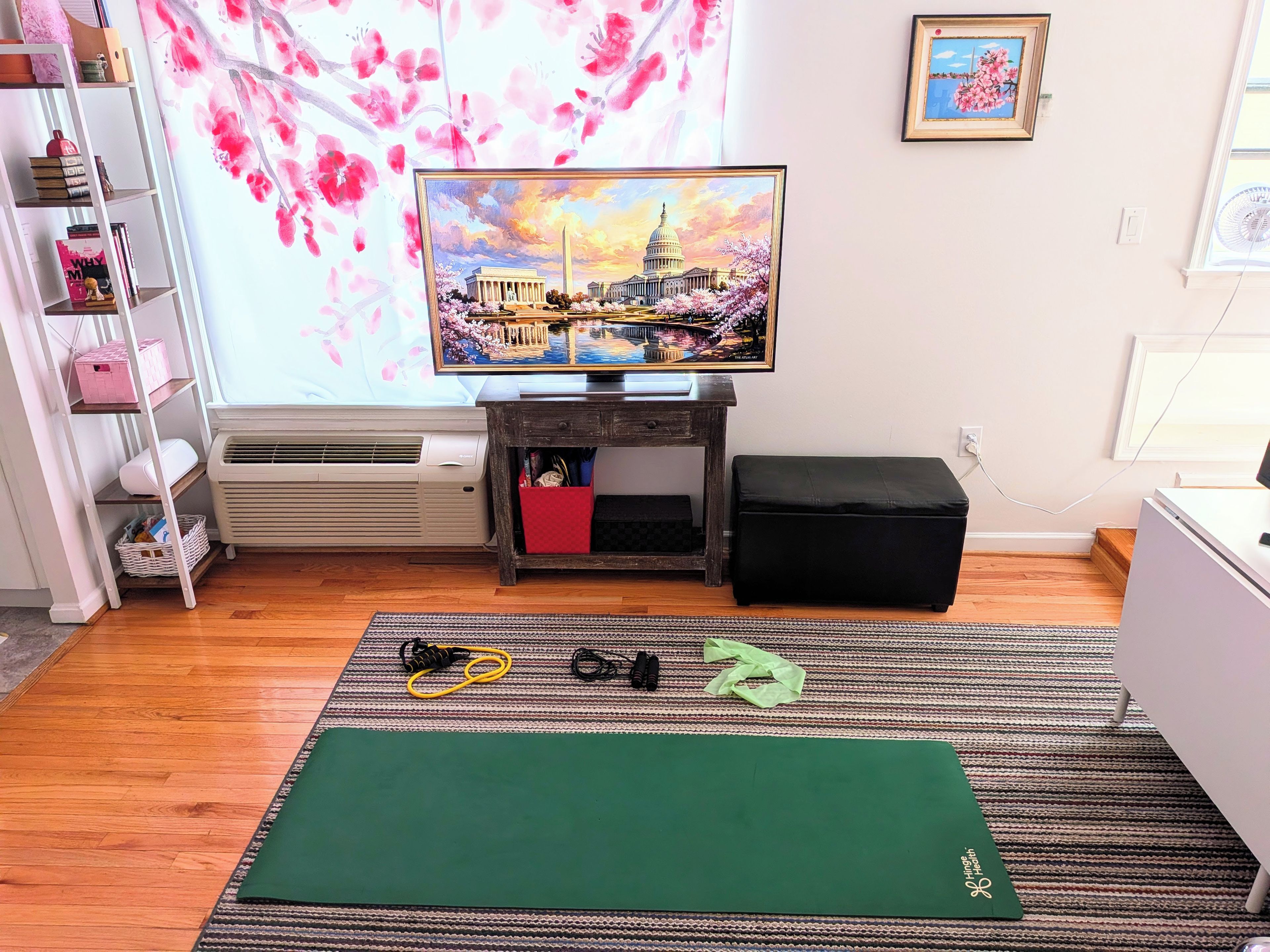 A room with a television on a stand, a bookshelf, a small table with a red basket, a black ottoman, and a green exercise mat.