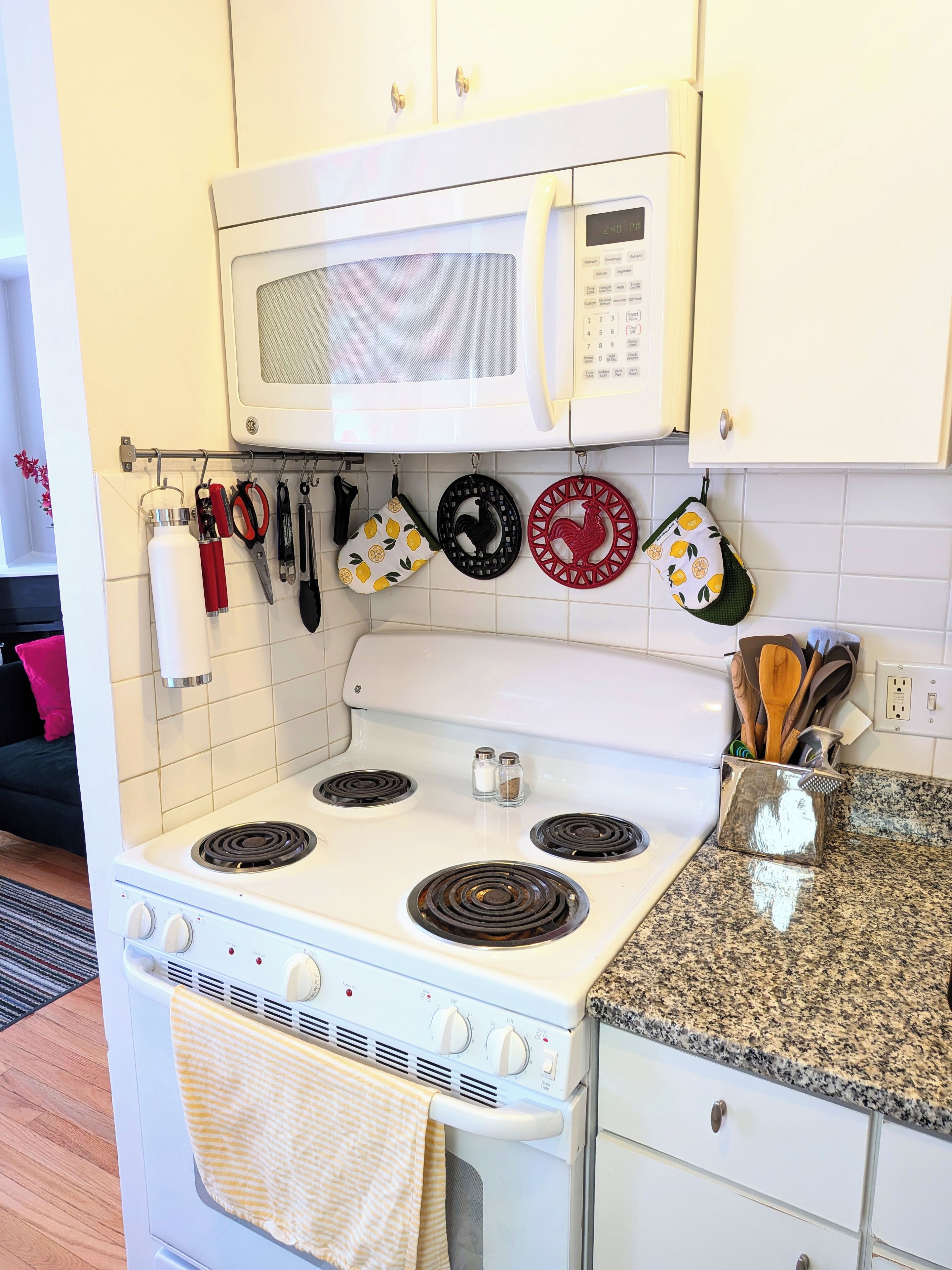A kitchen with a white stove, oven, and microwave, a granite countertop, and a hanging rack with kitchen utensils.