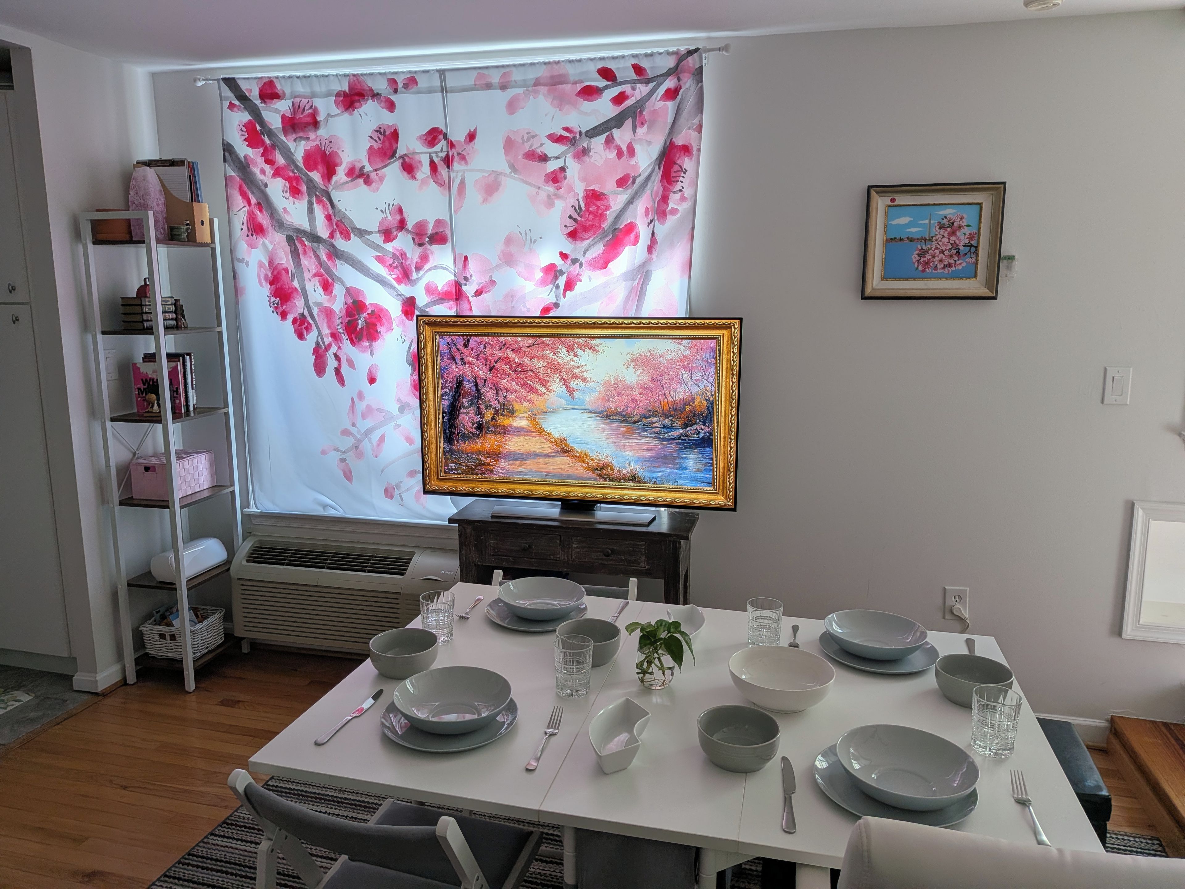 A dining area with a table set for four, a television displaying a landscape, and a bookshelf with various items.