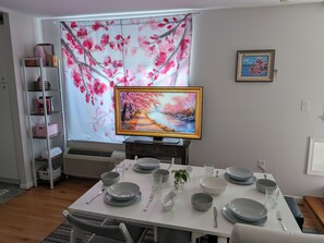 A dining area with a table set for four, a television displaying a landscape, and a bookshelf with various items.