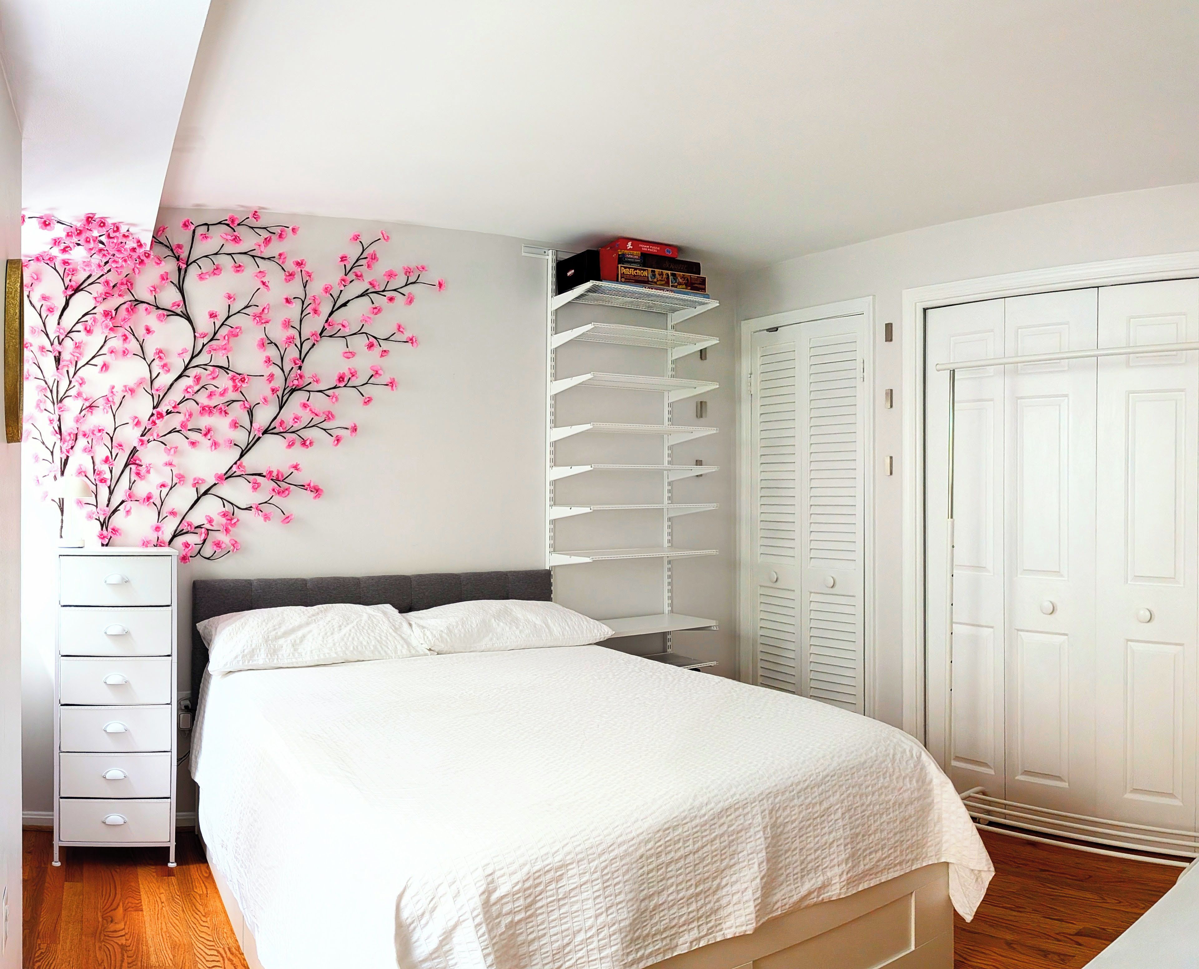 A bedroom with a bed, a dresser, a white wardrobe, and a shelving unit against the wall.