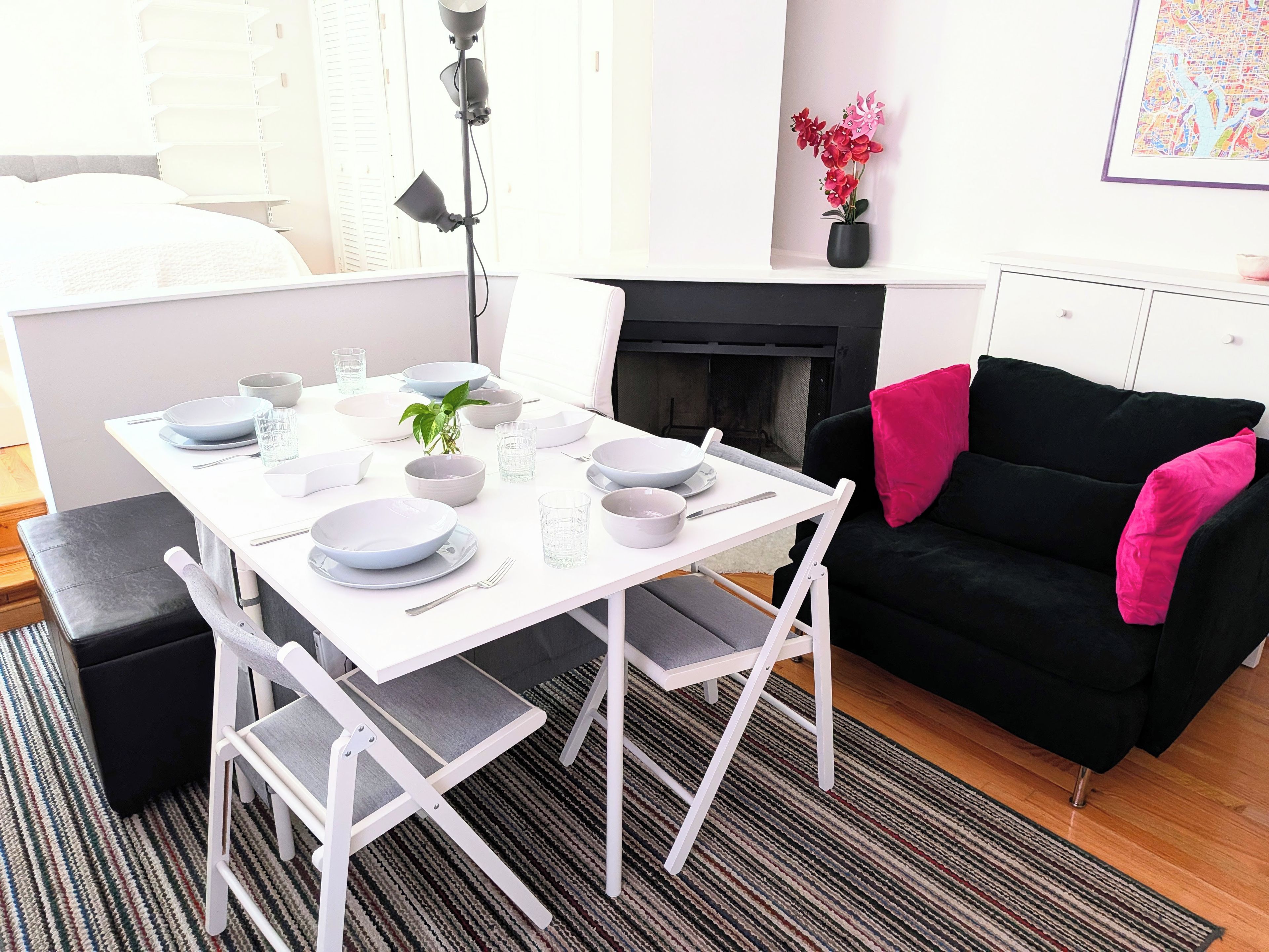A modern living room with a white dining table set for four, a black sofa with pink cushions, a fireplace, and a framed map on the wall.