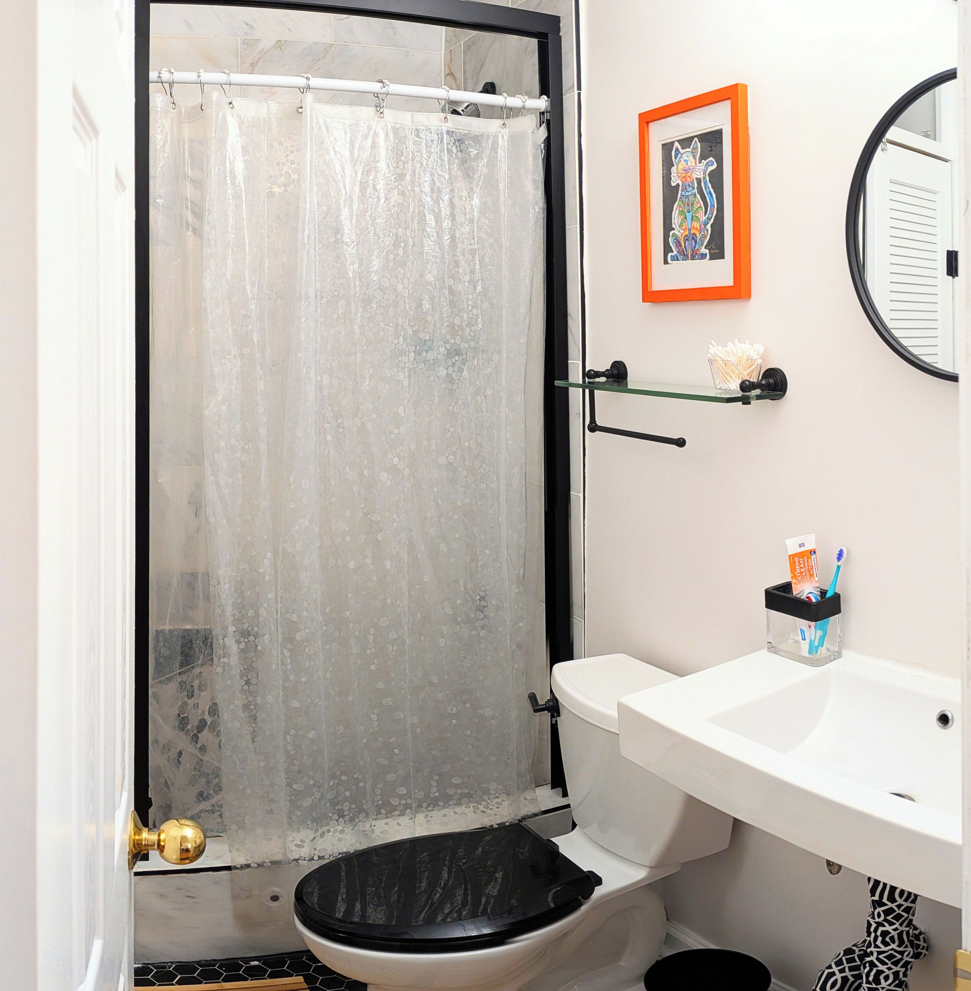 Ein Badezimmer mit Toilette, Waschbecken, Duschvorhang, einem gerahmten Bild an der Wand und einem kleinen Ablagefach für Toilettenartikel.