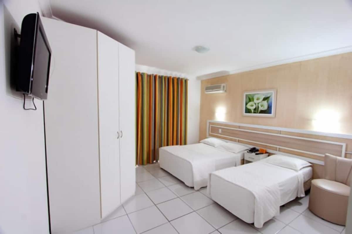 Quarto de hotel com duas camas, televisão, guarda-roupa, mesa de cabeceira, cadeira e janela com cortinas.