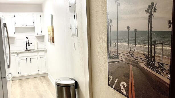 A kitchen with white cabinets, a stainless steel trash can, and a framed beach scene on the wall.