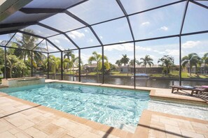 An indoor swimming pool with clear water, surrounded by a glass enclosure, and a view of a residential area with palm trees and houses. - Villa Coral Sunrise (Cape Coral)