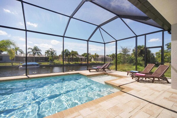 A swimming pool with a glass enclosure, two lounge chairs, and a view of a water body and greenery. - Villa Coral Sunrise (Cape Coral)