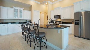 Una cocina con armarios blancos, una isla central con taburetes de bar, un refrigerador y un microondas.