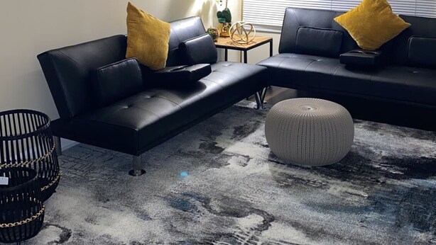 Un salon comprenant un canapé d’angle, un pouf rond, un tapis noir et blanc, un ventilateur de plafond et un tableau abstrait accroché au mur.