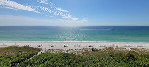 Uma praia com fileiras de espreguiçadeiras e guarda-sóis, céu claro e mar calmo.
