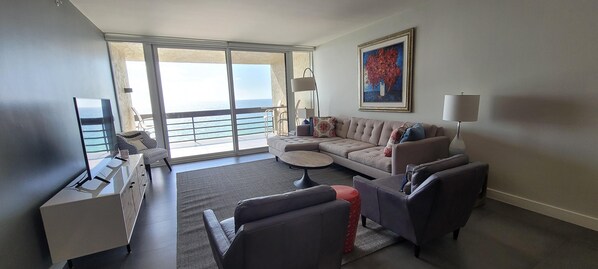 Uma sala de estar moderna com sofá, poltronas, mesa de centro e televisão. Há uma varanda com vista para o mar.