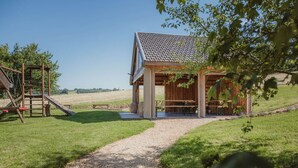 A rustic wooden structure with a tiled roof, a gravel path leading to it, and a playground area with a slide and swings. - La Bushaye - Cottage L'Etable (Aubel)