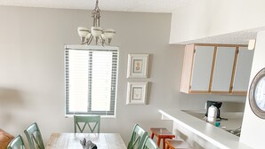 Ein Essbereich mit Holztisch und Stühlen, ein Fenster mit Jalousien, eine Küchenzeile mit Schränken und Spüle sowie eine an der Wand befestigte Uhr.