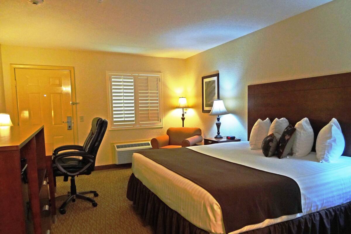 Une chambre d’hôtel comprenant un lit, un bureau, une chaise, une fenêtre avec des stores, une lampe et une horloge fixée au mur.