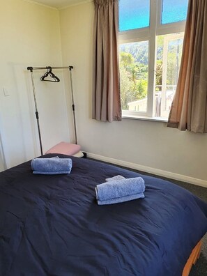 Un lit simple avec une couvre-lit bleu foncé, deux serviettes pliées, un oreiller rose et un cintre noir sur un porte-manteau fixé au mur.
