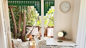 A room with a white table and chairs, a clock on the wall, and a view of a garden with trees and a fence. - Rêves de Soleil: Sweet Sunset Lodge with panoramic sea view (Sainte-Anne)