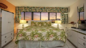 A bedroom with a floral bedspread, two bedside lamps, a large window with a sunset view, a television on a dresser, and a cabinet.