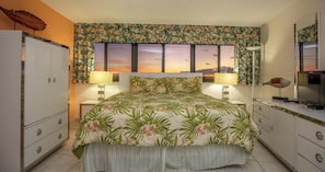 Un dormitorio con colcha estampada, dos lámparas de noche, un ventanal con vistas a un atardecer, un televisor sobre una cómoda y un armario.