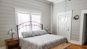 A bedroom with a bed, two bedside lamps, a wooden bench, a nightstand, a door, and a window with blinds. - Country house minutes from Aggieland  (College Station)