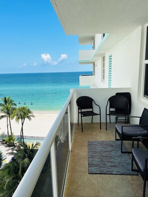 A balcony with a view of the ocean, two chairs, a small rug, and a window with a blue shutter.