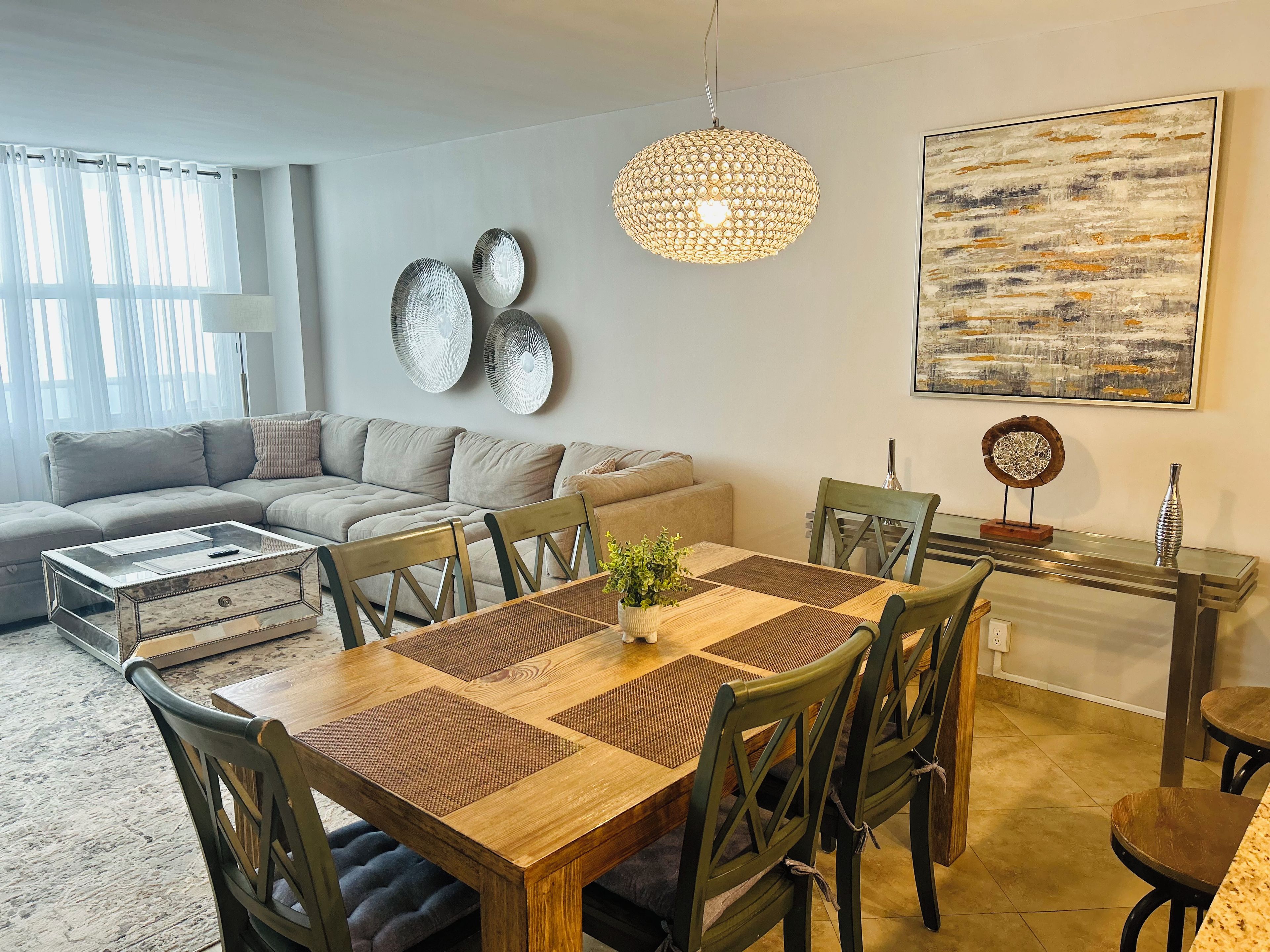 A modern living room with a dining area, featuring a sofa, a wooden dining table with chairs, a hanging light fixture, and a wall-mounted abstract painting.