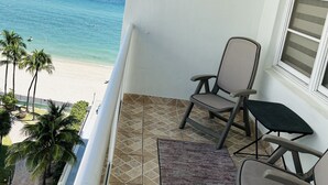 A balcony with two chairs and a small table, overlooking a beach with palm trees and the ocean.