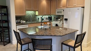 Una cocina con armarios de madera, encimera de granito, horno integrado, microondas y refrigerador. Hay dos sillas en la isla.