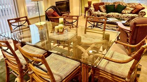 Una sala de estar con una mesa de comedor de vidrio rodeada de sillas de mimbre, un sofá con cojines estampados, una chimenea y un televisor.