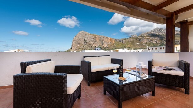 A terrace with wicker furniture, a glass table, and a view of a mountain and buildings.
