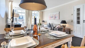 A dining area with a wooden table set for four, a large black pendant light, and a view of the city through large windows. - Residence Bismarck, Whg. 9 - Residence Bismarck (Sylt)