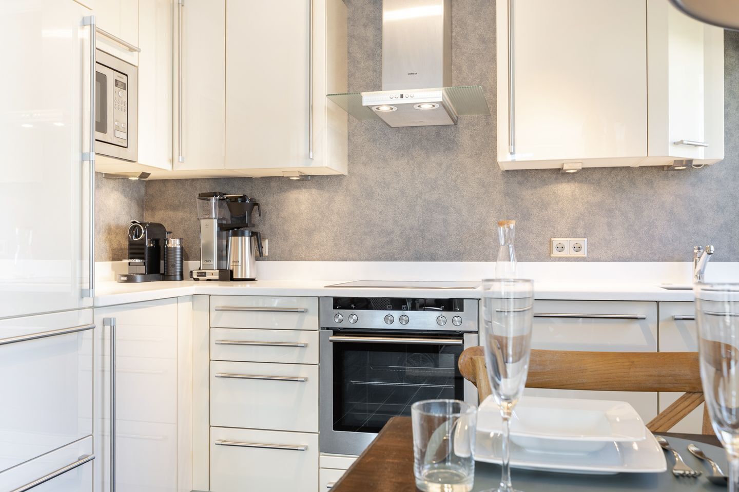 A modern kitchen with white cabinets, a built-in oven, a coffee machine, and a glass of water on the counter.